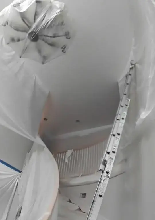Two-story foyer and curved staircase fully masked in plastic sheeting with a ladder in place, prepped for interior spray painting